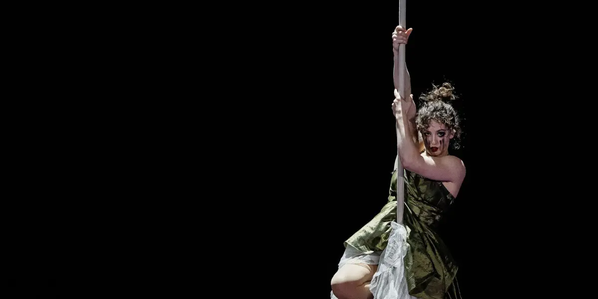 Une femme vêtue d'une robe verte et blanche effectue des acrobaties aériennes, éclairée sur un fond sombre. Flavie Beaulieu se suspend gracieusement et prend la pose sur une corde, avec une corde enroulée reposant sur le sol en dessous.