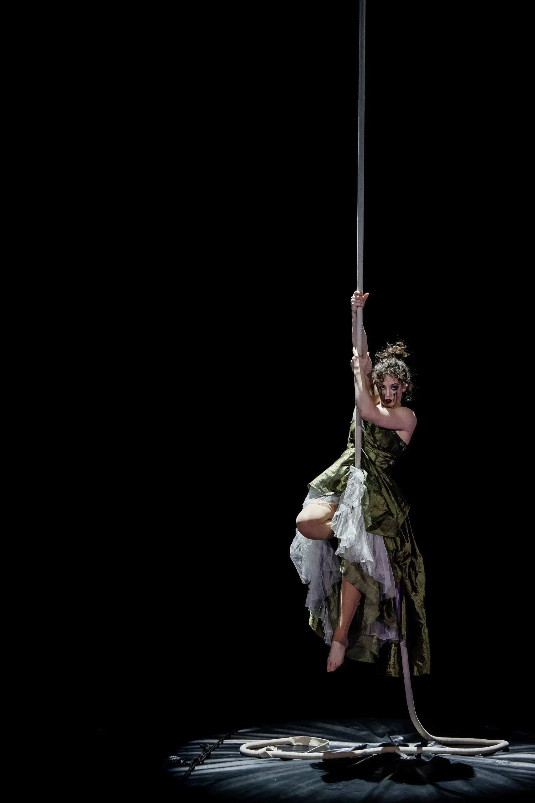 Une femme vêtue d'une robe verte et blanche effectue des acrobaties aériennes, éclairée sur un fond sombre. Flavie Beaulieu se suspend gracieusement et prend la pose sur une corde, avec une corde enroulée reposant sur le sol en dessous.