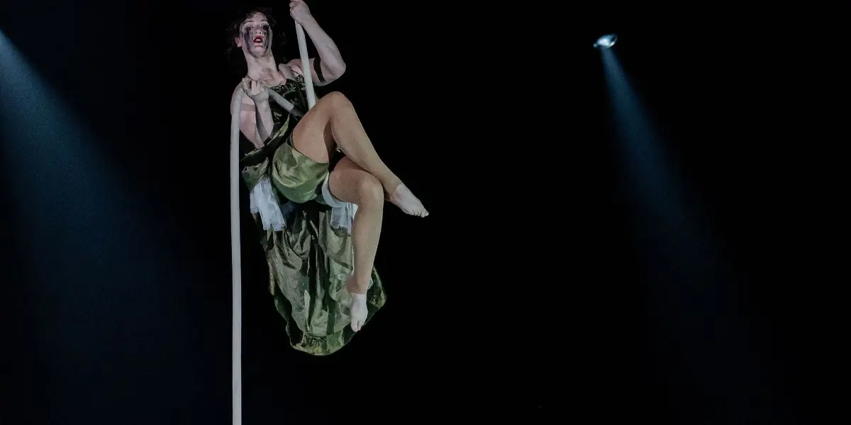 L'artiste aérienne Flavie Beaulieu, vêtue d'un costume vert, est suspendue à une corde blanche au-dessus de la scène, souriante, tandis que des projecteurs spectaculaires éclairent l'arrière-plan sombre.