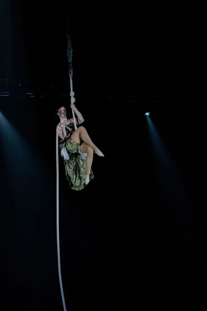 L'artiste aérienne Flavie Beaulieu, vêtue d'un costume vert, est suspendue à une corde blanche au-dessus de la scène, souriante, tandis que des projecteurs spectaculaires éclairent l'arrière-plan sombre.