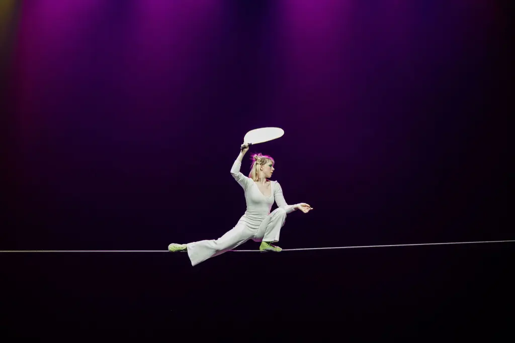 Hannah Grove, vêtue d'une tenue blanche et de chaussures vertes, se tient en équilibre sur une corde raide tout en tenant une pagaie blanche au-dessus de sa tête, sur fond de lumière violette.