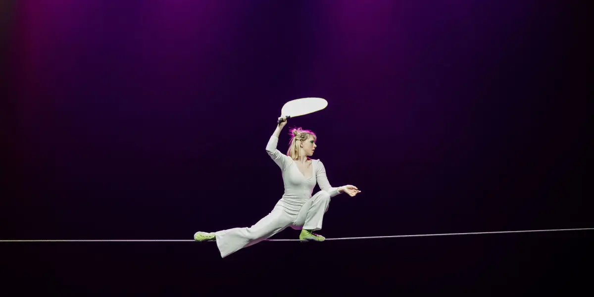 Hannah Grove, vêtue d'une tenue blanche et de chaussures vertes, se tient en équilibre sur une corde raide tout en tenant une pagaie blanche au-dessus de sa tête, sur fond de lumière violette.