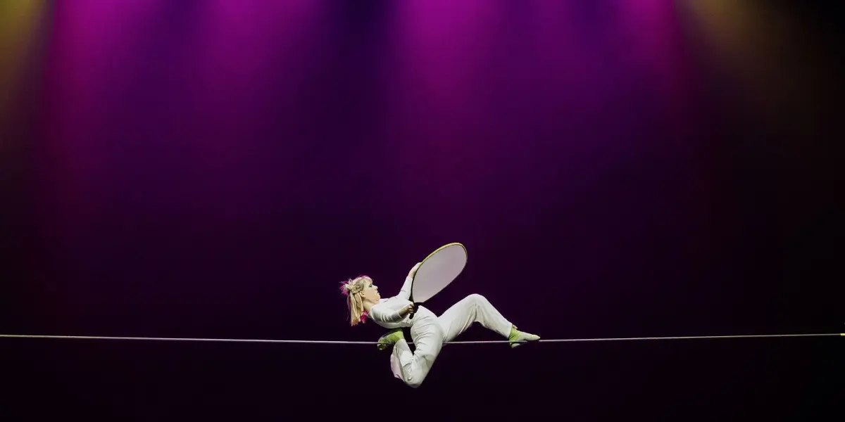 Un artiste vêtu d'un costume blanc et de chaussures vertes se tient en équilibre sur une corde raide tout en tenant un accessoire circulaire, sous un éclairage dramatique violet et jaune - un acte qui rappelle le style captivant d'Hannah Grove.