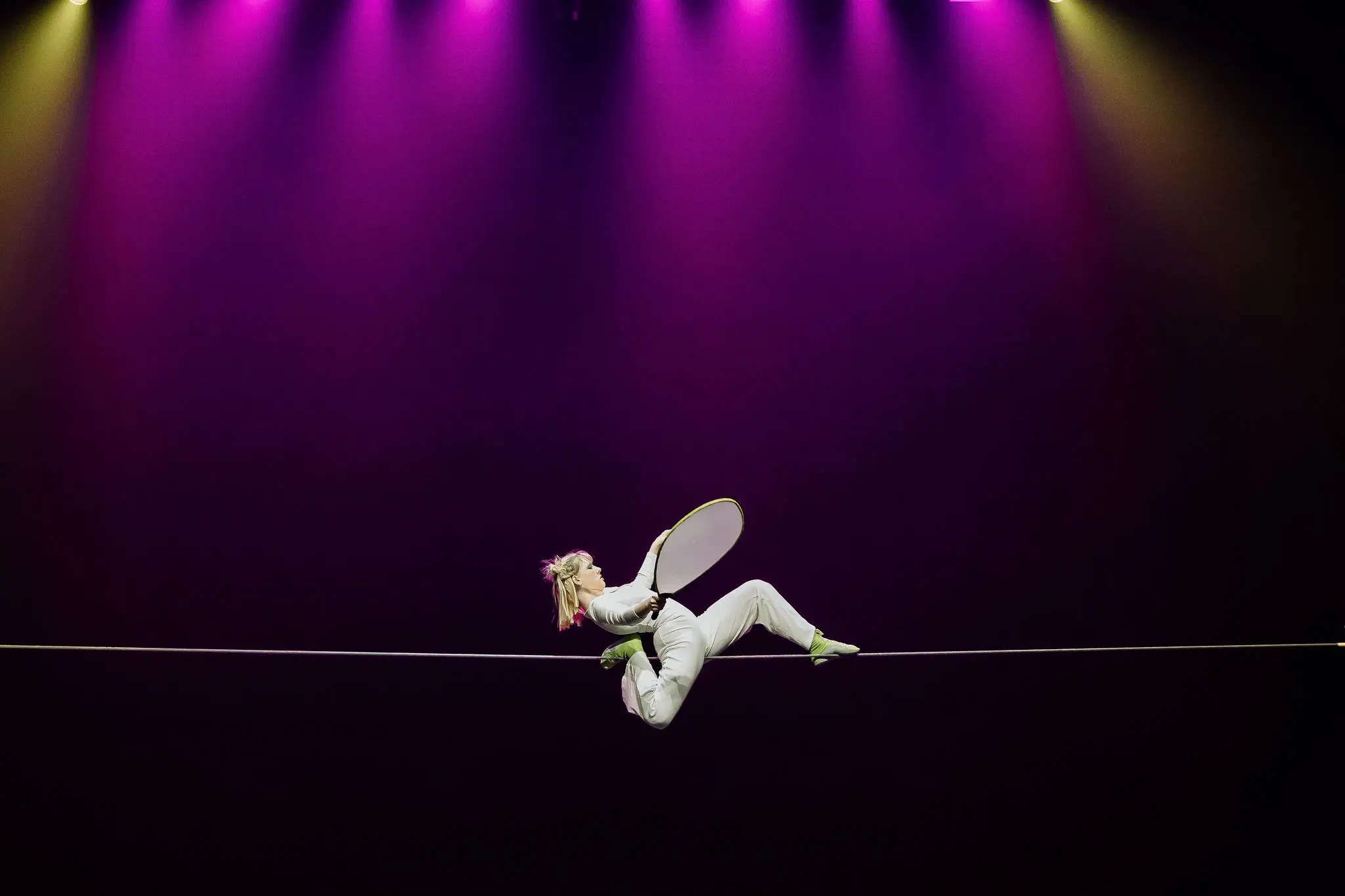 Un artiste vêtu d'un costume blanc et de chaussures vertes se tient en équilibre sur une corde raide tout en tenant un accessoire circulaire, sous un éclairage dramatique violet et jaune - un acte qui rappelle le style captivant d'Hannah Grove.
