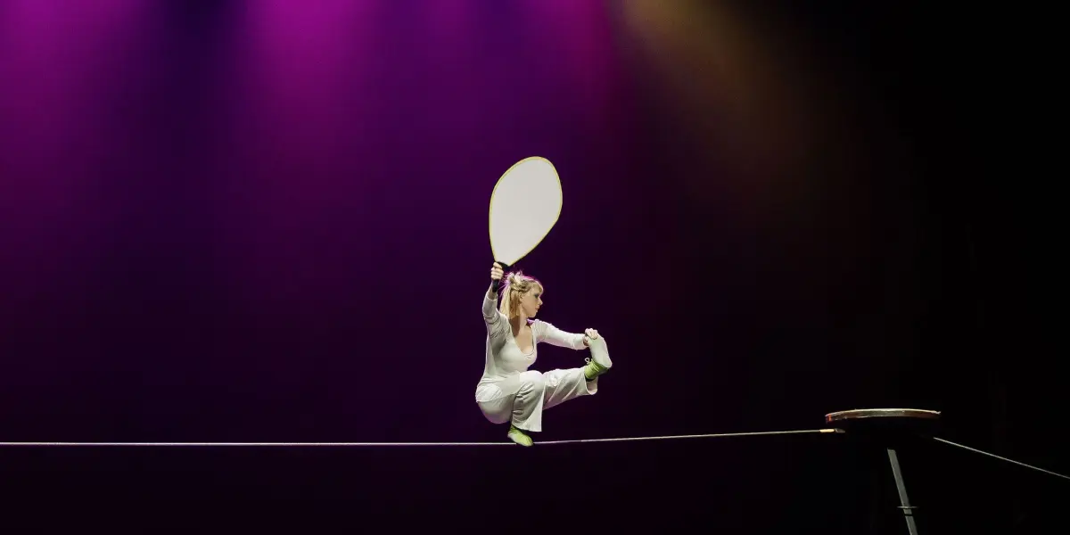 Hannah Grove, vêtue de blanc, se tient en équilibre sur une corde raide sous des lumières de scène violettes et jaunes, tenant une grande pagaie tout en étant assise avec une jambe tendue et une jambe pliée.