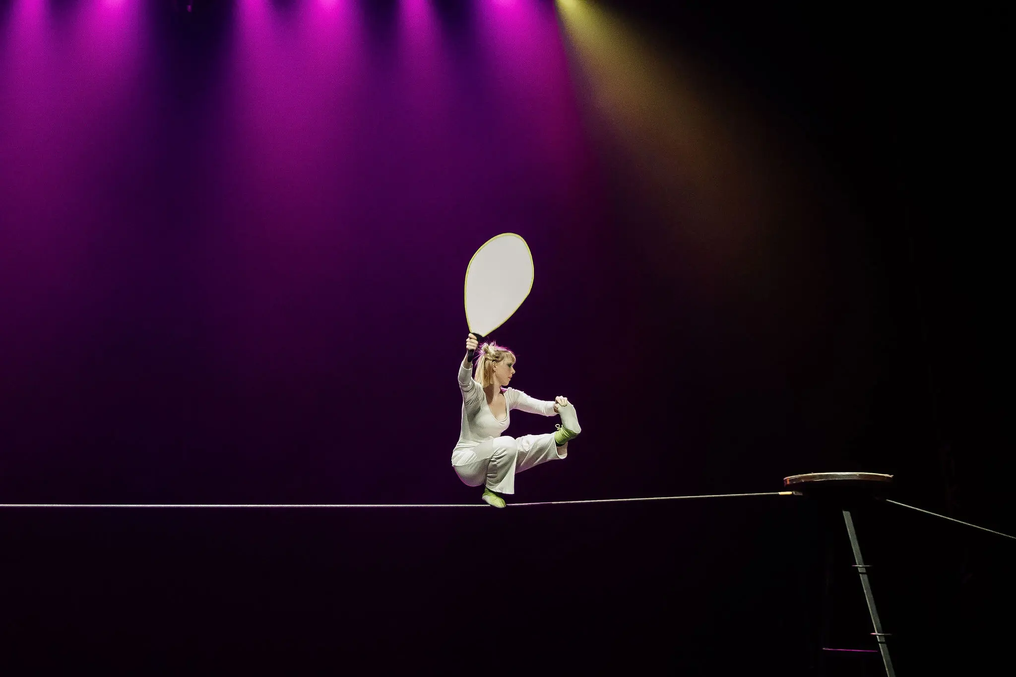 Hannah Grove, vêtue de blanc, se tient en équilibre sur une corde raide sous des lumières de scène violettes et jaunes, tenant une grande pagaie tout en étant assise avec une jambe tendue et une jambe pliée.