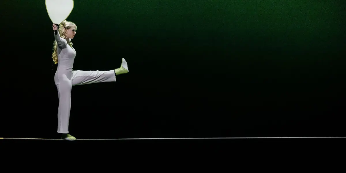 Hannah Grove, en tenue claire, se tient en équilibre sur une corde raide, levant une jambe et tenant une grande pagaie blanche sur un fond vert foncé.