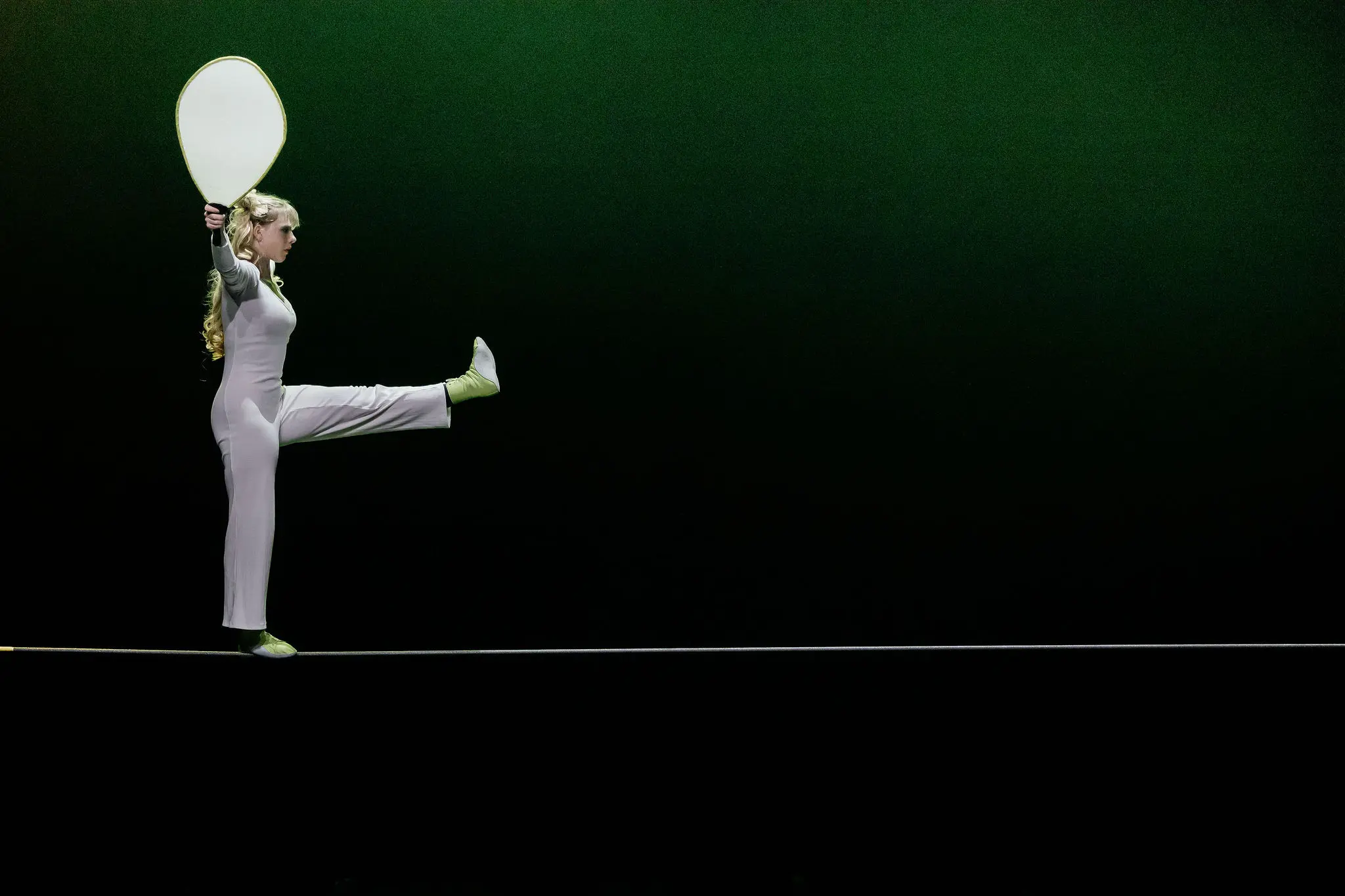 Hannah Grove, en tenue claire, se tient en équilibre sur une corde raide, levant une jambe et tenant une grande pagaie blanche sur un fond vert foncé.