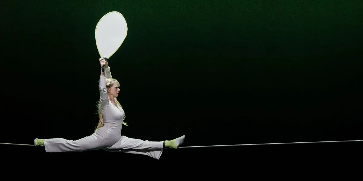 Hannah Grove, en tenue légère, effectue un grand écart sur une corde raide, tenant une grande pagaie blanche au-dessus de sa tête sur un fond vert foncé.