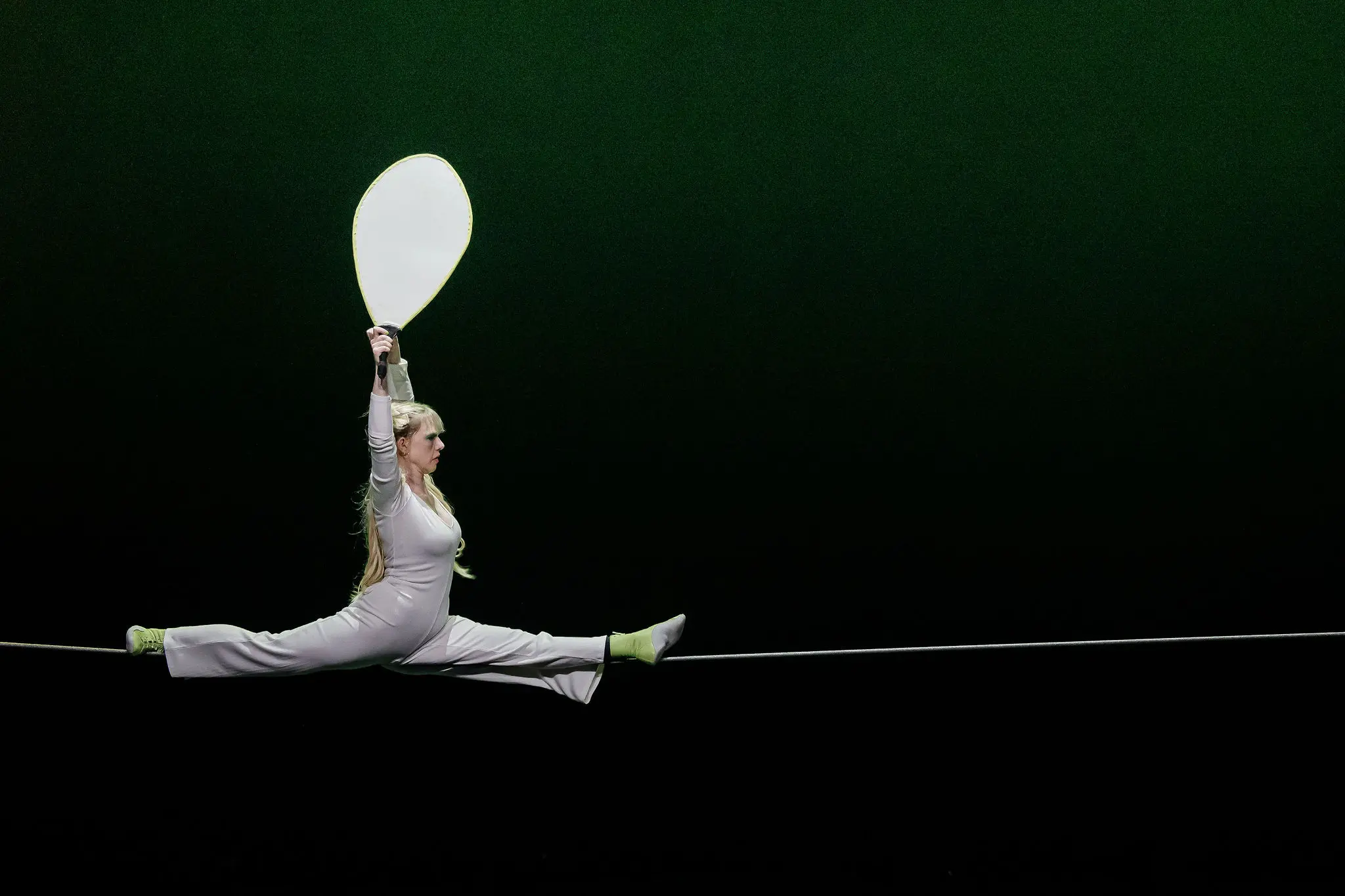 Hannah Grove, en tenue légère, effectue un grand écart sur une corde raide, tenant une grande pagaie blanche au-dessus de sa tête sur un fond vert foncé.