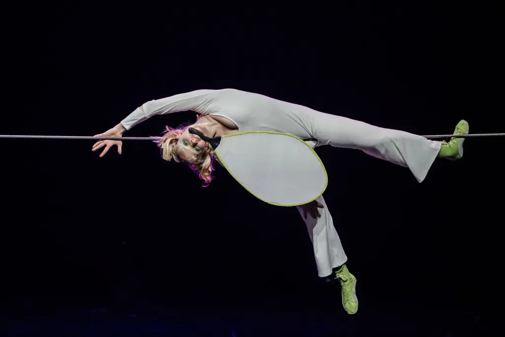 Hannah Grove, vêtue d'un costume blanc et de chaussures vertes fluo, se tient en équilibre sur une corde raide, le corps arqué vers l'arrière, une main agrippée à la corde, sur un fond sombre.