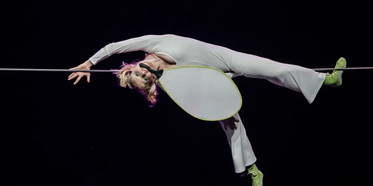 Hannah Grove, vêtue d'un costume blanc et de chaussures vertes fluo, se tient en équilibre sur une corde raide, le corps arqué vers l'arrière, une main agrippée à la corde, sur un fond sombre.