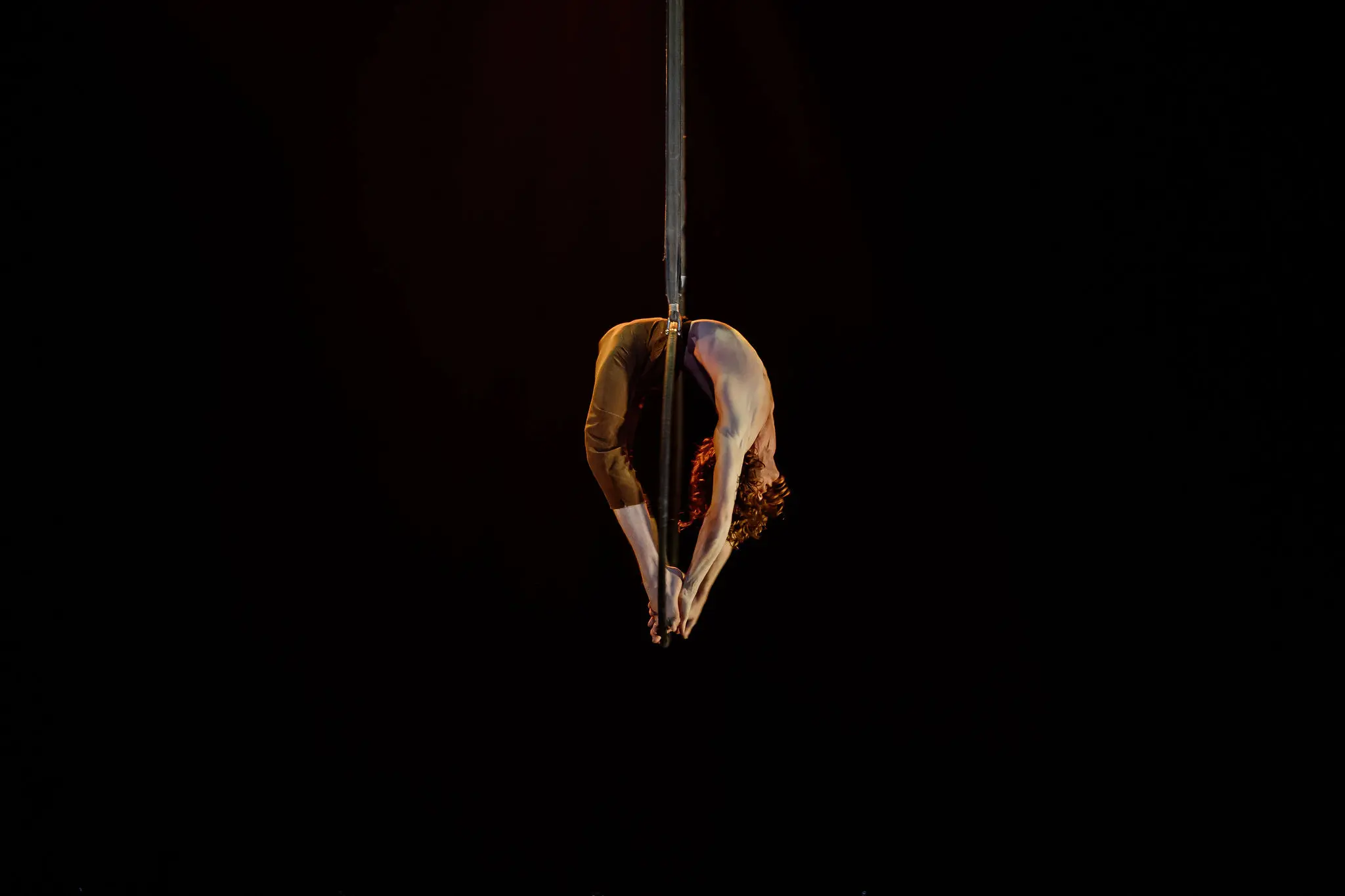 L'artiste aérien Jean Roubieu est suspendu à l'envers sur une corde, se penchant vers l'arrière pour toucher ses pieds, avec un arrière-plan sombre mettant en valeur sa pose.