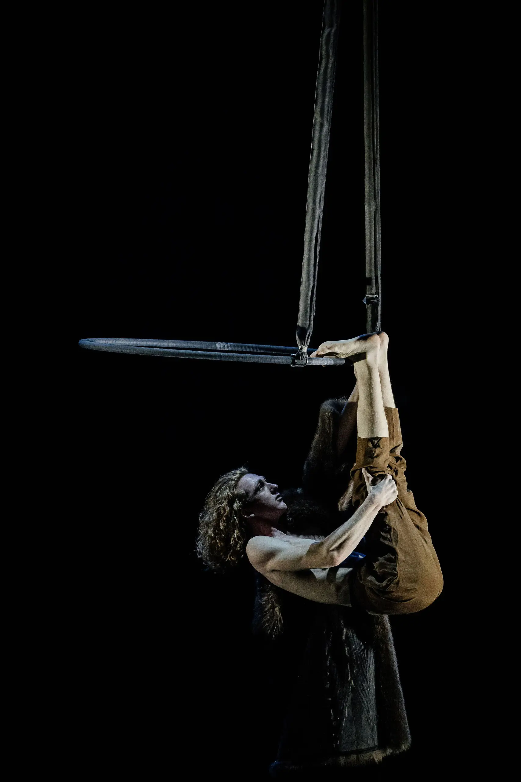 L'artiste aérien Jean Roubieu, cheveux bouclés et pantalon marron, est suspendu à l'envers à des sangles, s'agrippant avec les pieds et les mains sur un fond sombre.