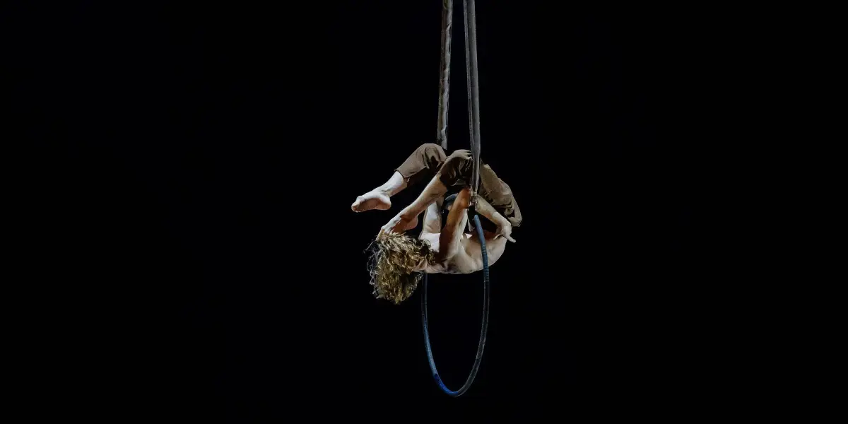 Un artiste de cirque aux cheveux bouclés est suspendu à l'envers à un cerceau aérien, les jambes et les bras enroulés autour du cerceau sur un fond noir et sombre, faisant écho à l'audace artistique de Jean Roubieu.
