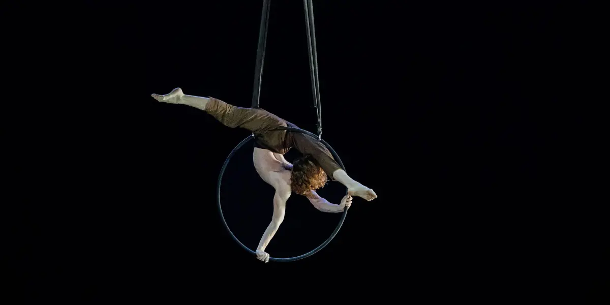 Un artiste de cirque aux cheveux bouclés est suspendu à l'envers à un cerceau aérien, étendant une jambe et un bras vers l'extérieur sur un fond sombre, rappelant l'art de Jean Roubieu.