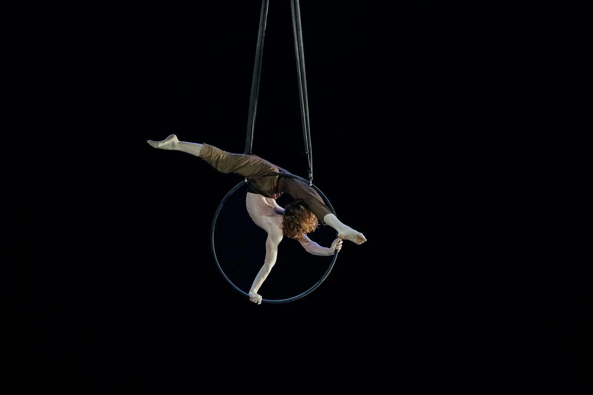 Un artiste de cirque aux cheveux bouclés est suspendu à l'envers à un cerceau aérien, étendant une jambe et un bras vers l'extérieur sur un fond sombre, rappelant l'art de Jean Roubieu.