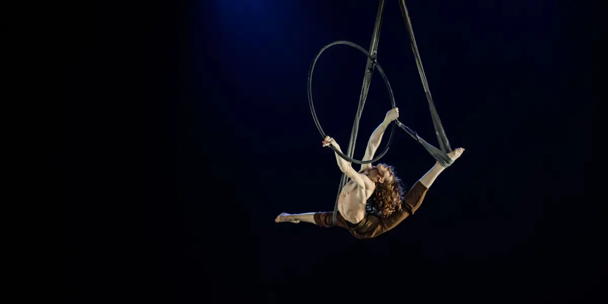Un acrobate aérien exécute un saut en suspension sur une scène sombre, éclairée par des projecteurs bleus, rappelant les performances captivantes souvent capturées par Jean Roubieu.