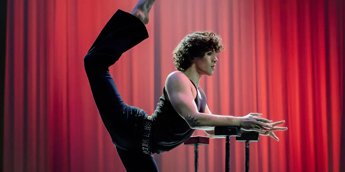 Cooper Yarosloski, un artiste souple, se tient en équilibre sur une jambe au sommet d'une plate-forme, l'autre jambe étant tendue loin derrière et les mains reposant sur des barres parallèles, sur un fond de scène éclairé par des lumières rouges et orange.