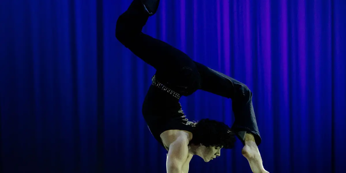 Cooper Yarosloski, vêtu d'un pantalon noir, effectue une impressionnante pose d'équilibre sur deux cannes contre un rideau bleu foncé éclairé par la scène. Le corps est arqué, les jambes gracieusement repliées au-dessus de la tête.