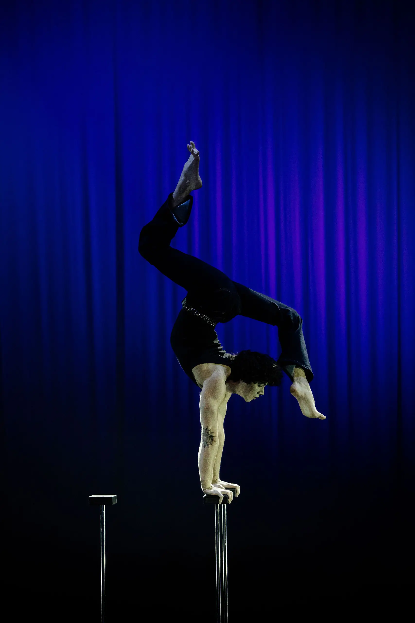 Cooper Yarosloski, vêtu d'un pantalon noir, effectue une impressionnante pose d'équilibre sur deux cannes contre un rideau bleu foncé éclairé par la scène. Le corps est arqué, les jambes gracieusement repliées au-dessus de la tête.