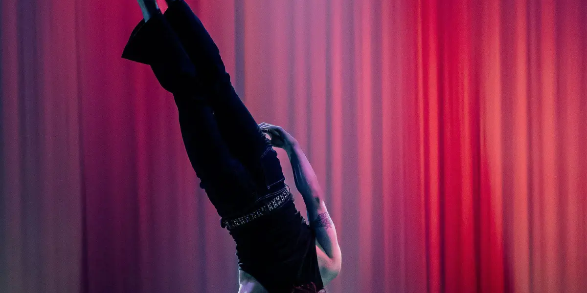 L'acrobate Cooper Yarosloski réalise un équilibre à une main sur un poteau, les jambes tendues d'un côté, sur un fond de scène avec un éclairage dramatique rouge et violet et des projecteurs en dessous.