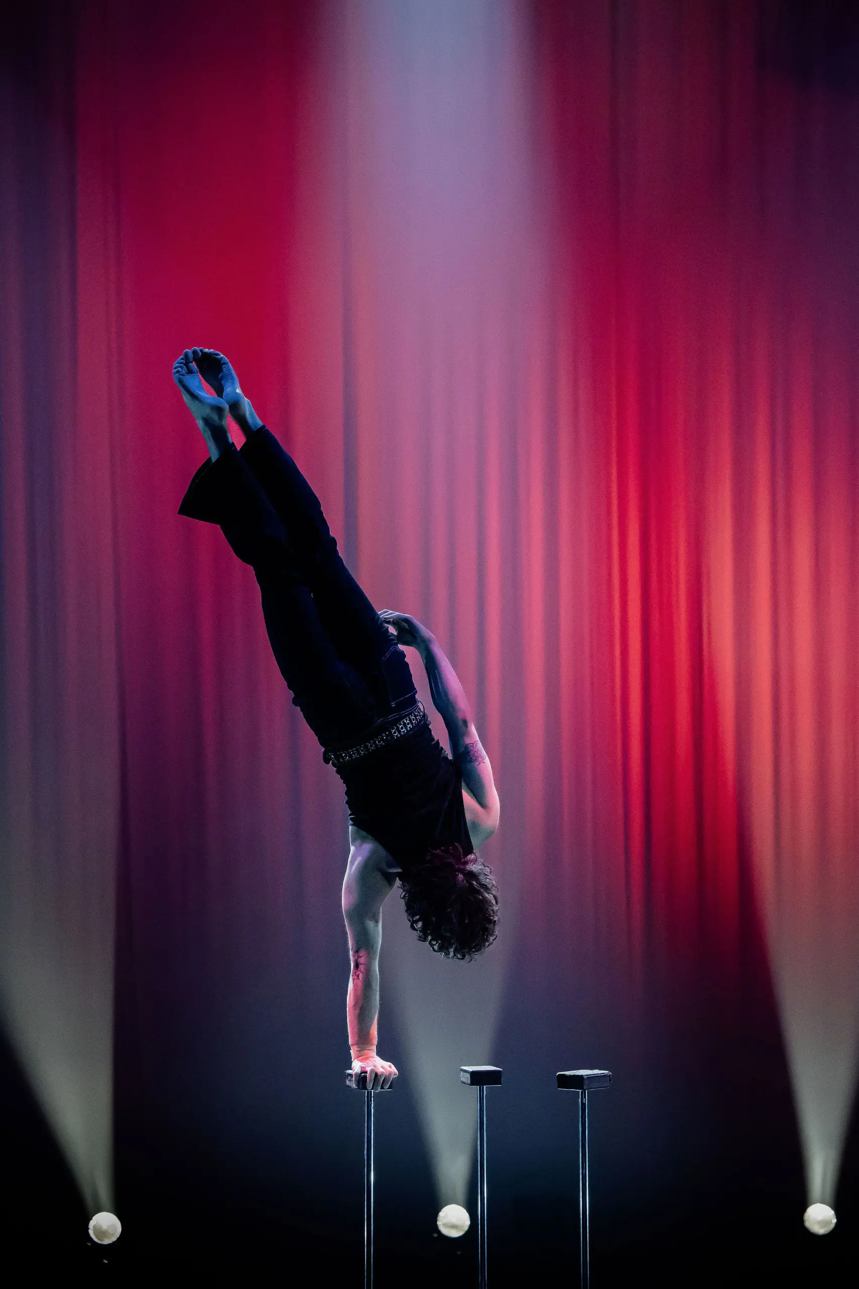 L'acrobate Cooper Yarosloski réalise un équilibre à une main sur un poteau, les jambes tendues d'un côté, sur un fond de scène avec un éclairage dramatique rouge et violet et des projecteurs en dessous.