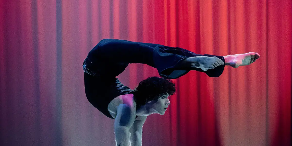 L'acrobate Cooper Yarosloski, torse nu, se tient en équilibre sur les mains au sommet de deux poteaux, le dos arqué et les jambes au-dessus de la tête, sur un fond de scène éclairé par des lumières rouges et des projecteurs.