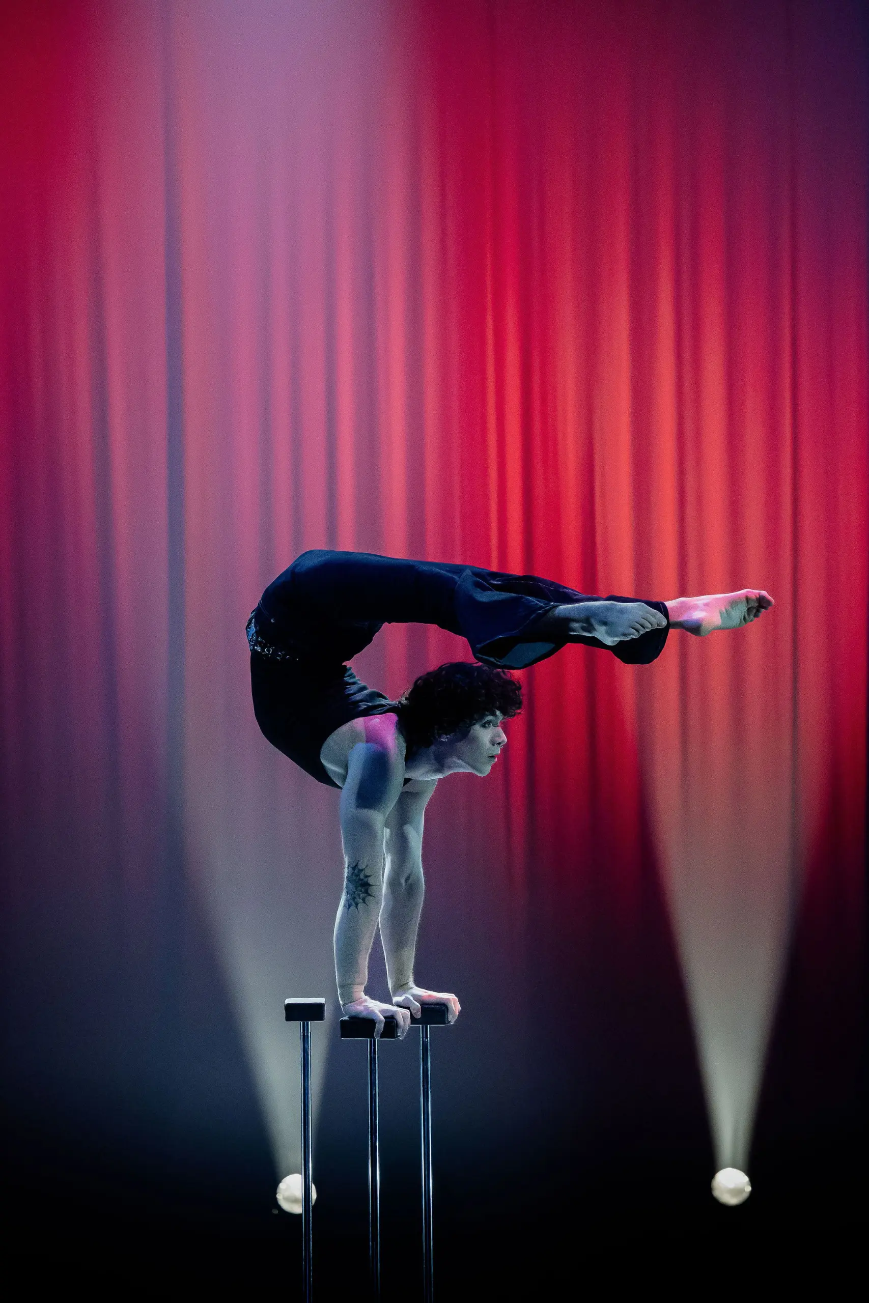 L'acrobate Cooper Yarosloski, torse nu, se tient en équilibre sur les mains au sommet de deux poteaux, le dos arqué et les jambes au-dessus de la tête, sur un fond de scène éclairé par des lumières rouges et des projecteurs.