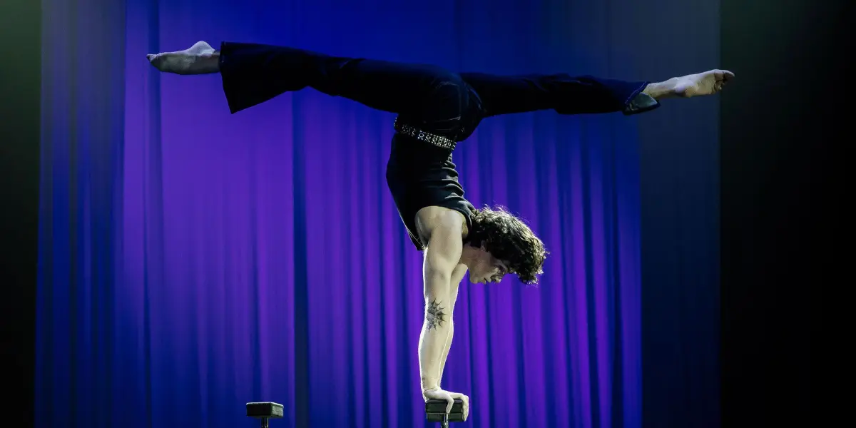 Cooper Yarosloski, un artiste de cirque, se tient en équilibre sur une main au sommet de grandes perches, les jambes tendues en fente, sur un fond de scène éclairé en violet.