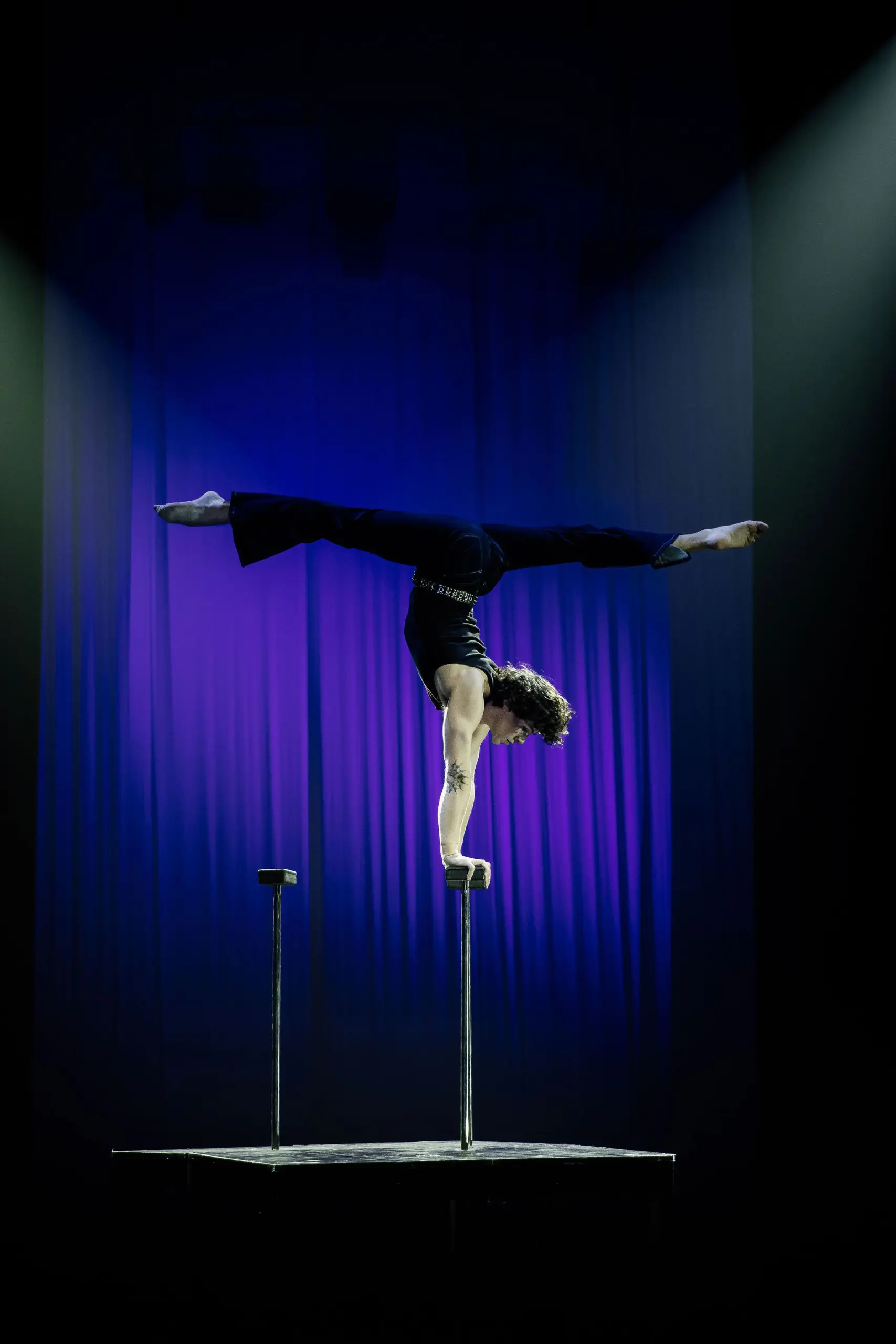 Cooper Yarosloski, un artiste de cirque, se tient en équilibre sur une main au sommet de grandes perches, les jambes tendues en fente, sur un fond de scène éclairé en violet.