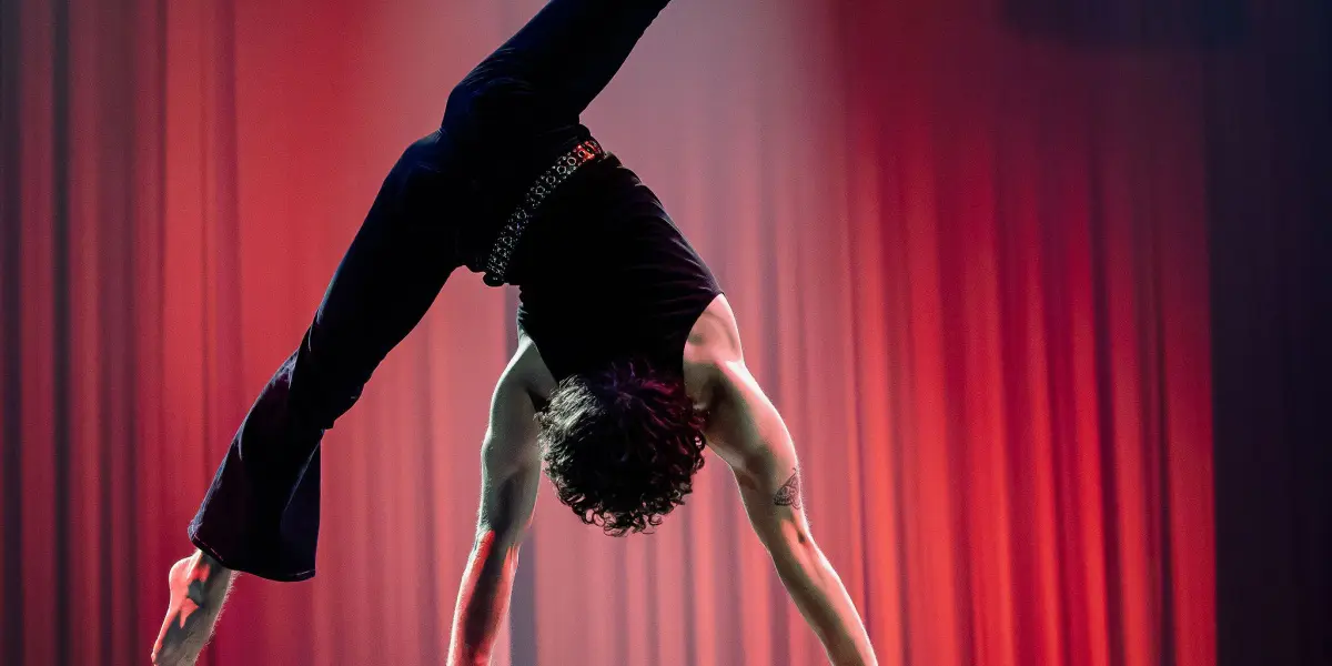 Cooper Yarosloski se tient en équilibre sur deux cannes, une jambe tendue et l'autre pliée, vêtu de noir sur une scène inondée d'un éclairage rouge dramatique et d'un projecteur.