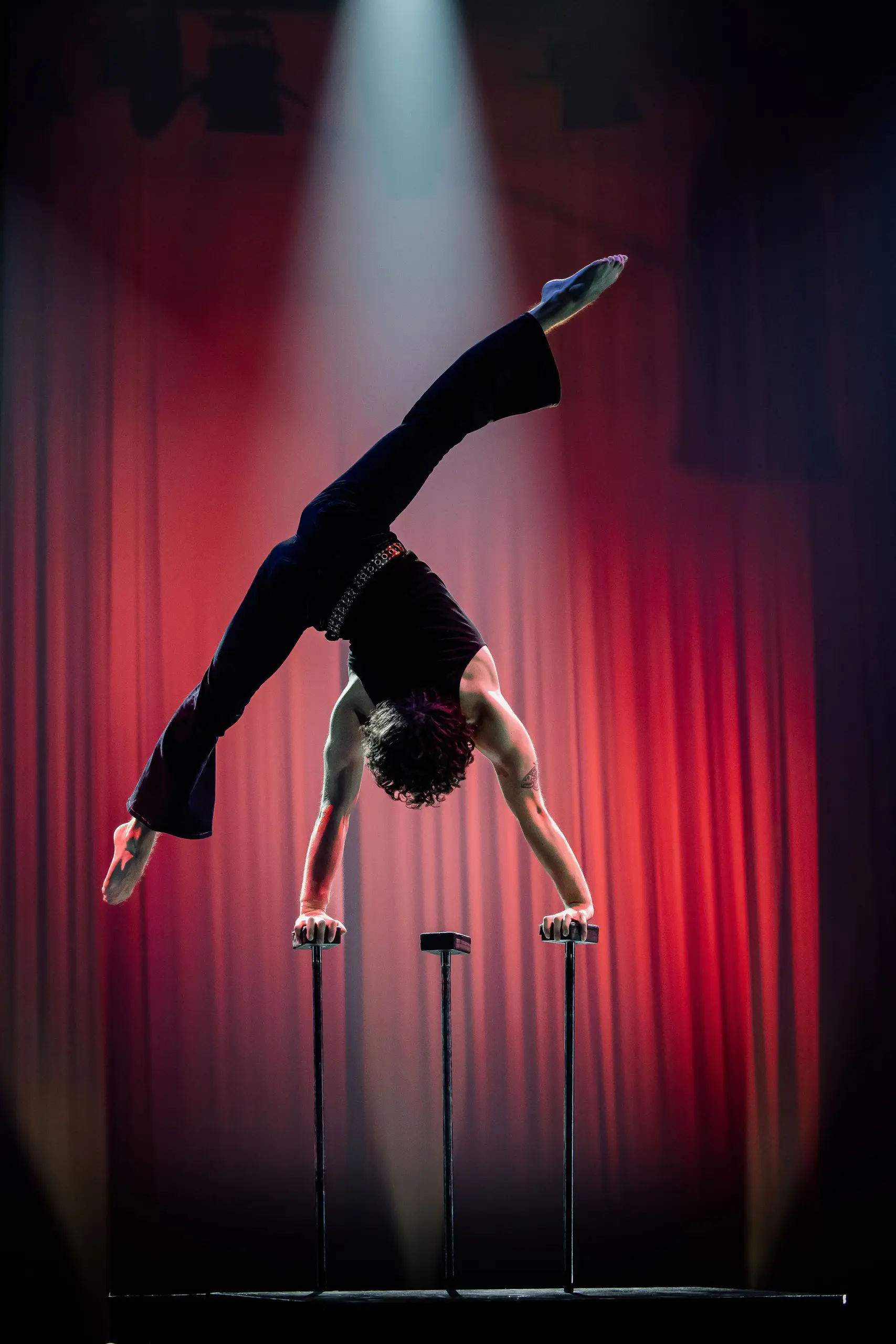 Cooper Yarosloski se tient en équilibre sur deux cannes, une jambe tendue et l'autre pliée, vêtu de noir sur une scène inondée d'un éclairage rouge dramatique et d'un projecteur.