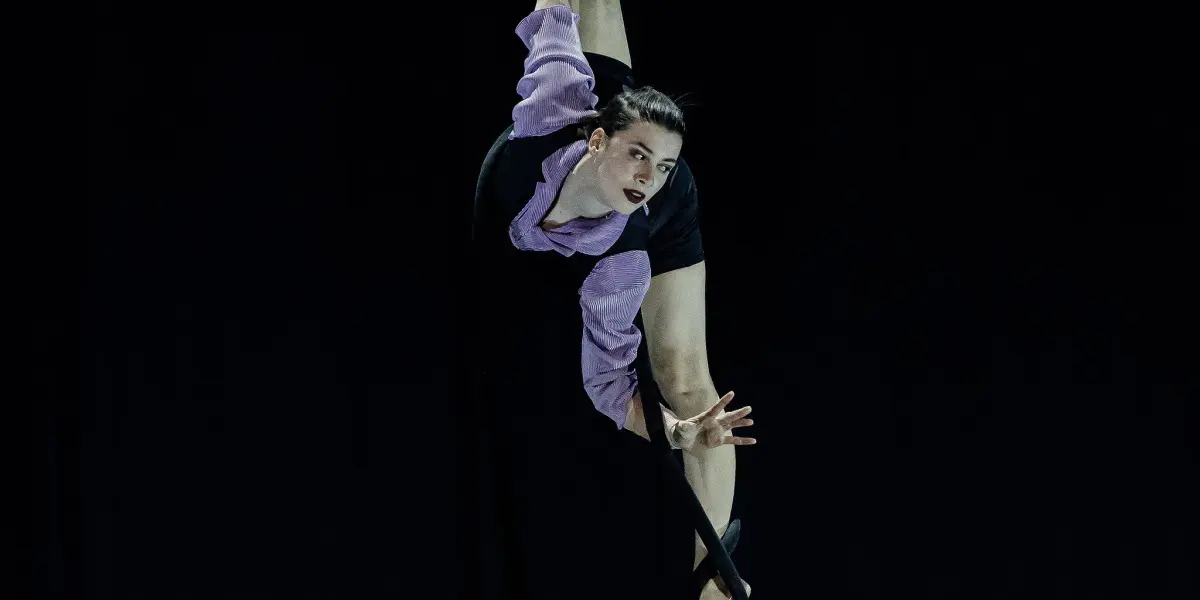L'acrobate aérienne Katarina Sobinkovicova, en tenue violette et noire, est suspendue à l'envers à une corde de soie noire, tendant une jambe vers le haut et étendant un bras avec grâce sur un fond sombre.