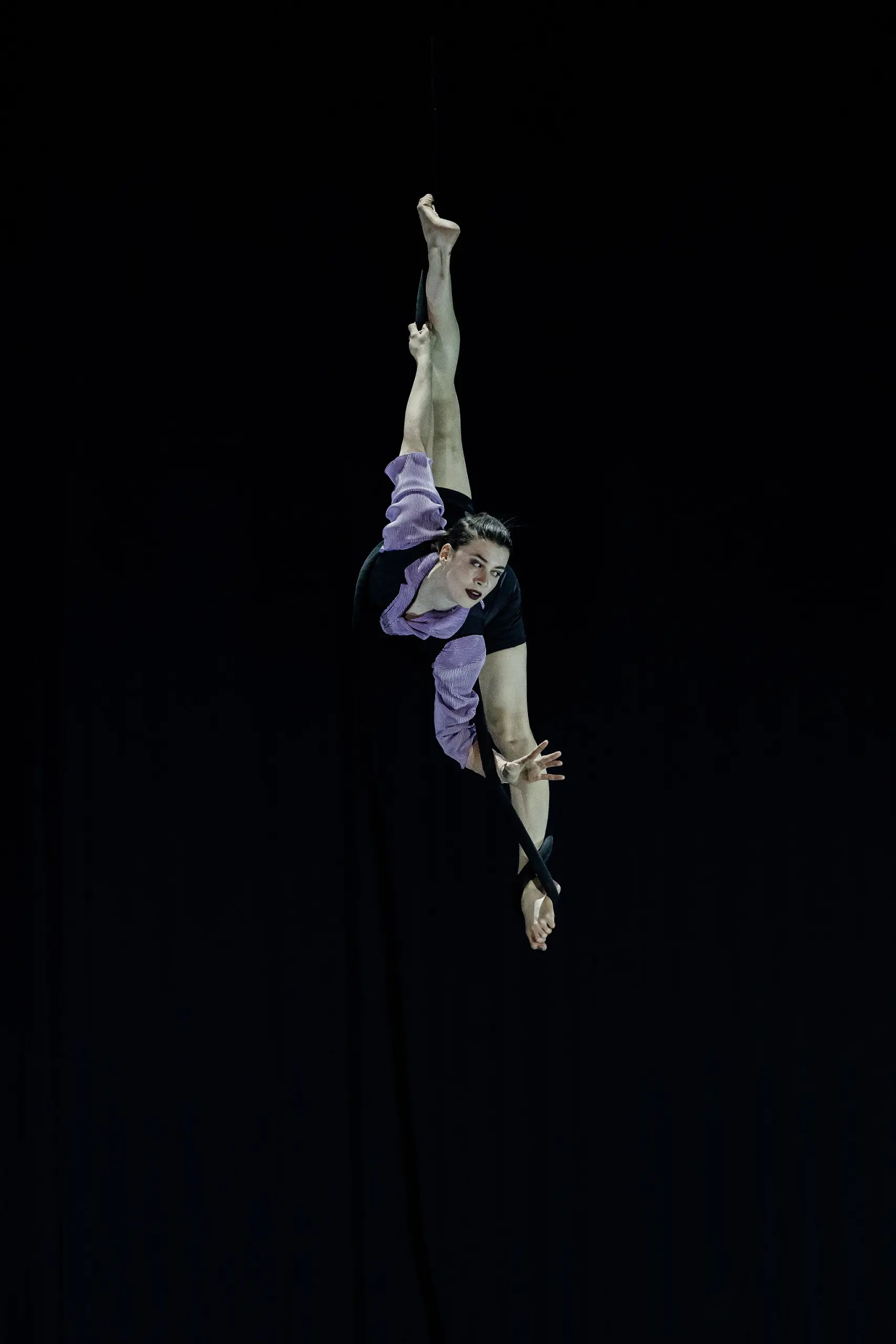L'acrobate aérienne Katarina Sobinkovicova, en tenue violette et noire, est suspendue à l'envers à une corde de soie noire, tendant une jambe vers le haut et étendant un bras avec grâce sur un fond sombre.