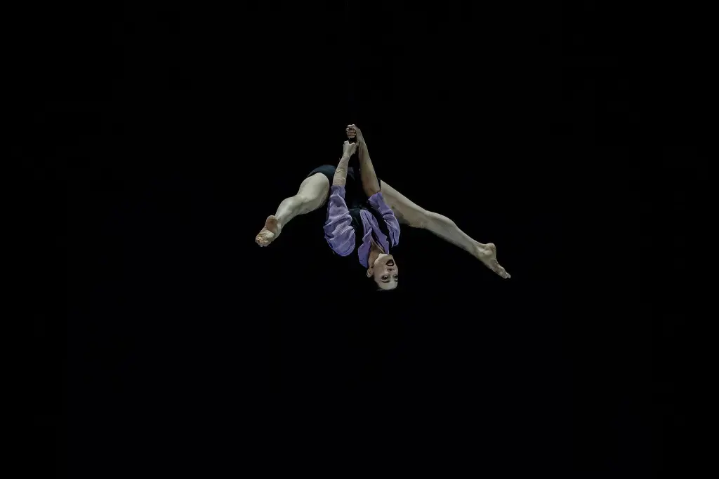L'artiste aérienne Katarina Sobinkovicova, vêtue d'une tenue violette, est suspendue la tête en bas, agrippée à une corde suspendue avec les deux mains et les jambes écartées, sur un fond sombre.