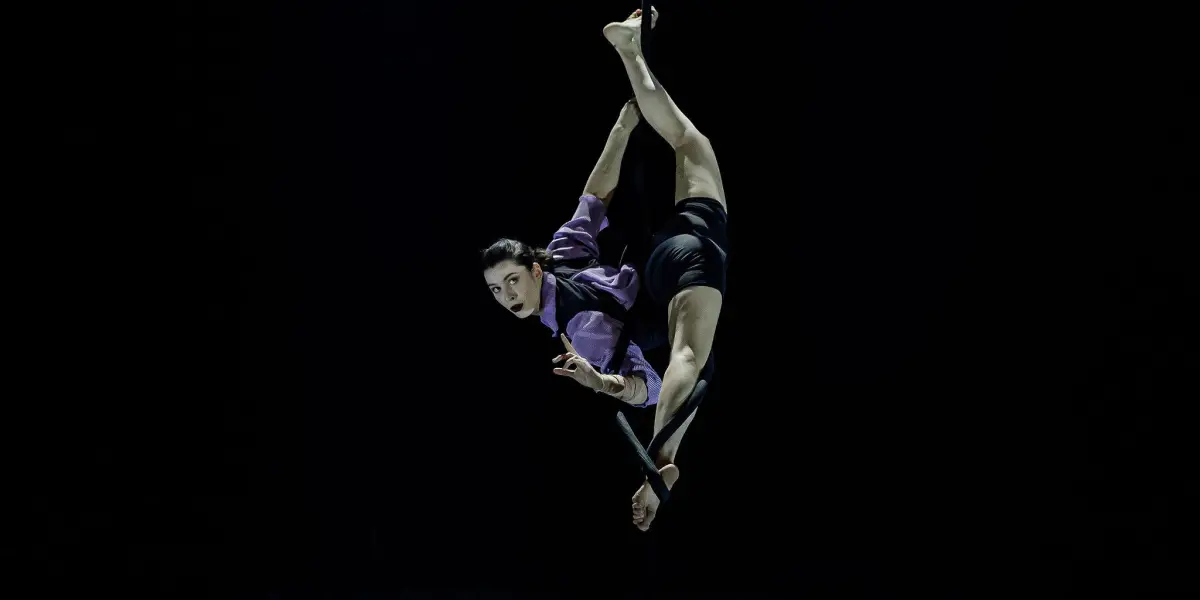 L'artiste aérienne Katarina Sobinkovicova se produit en hauteur, suspendue par une jambe à une corde, vêtue d'un haut violet et d'un short noir, sur un fond sombre.