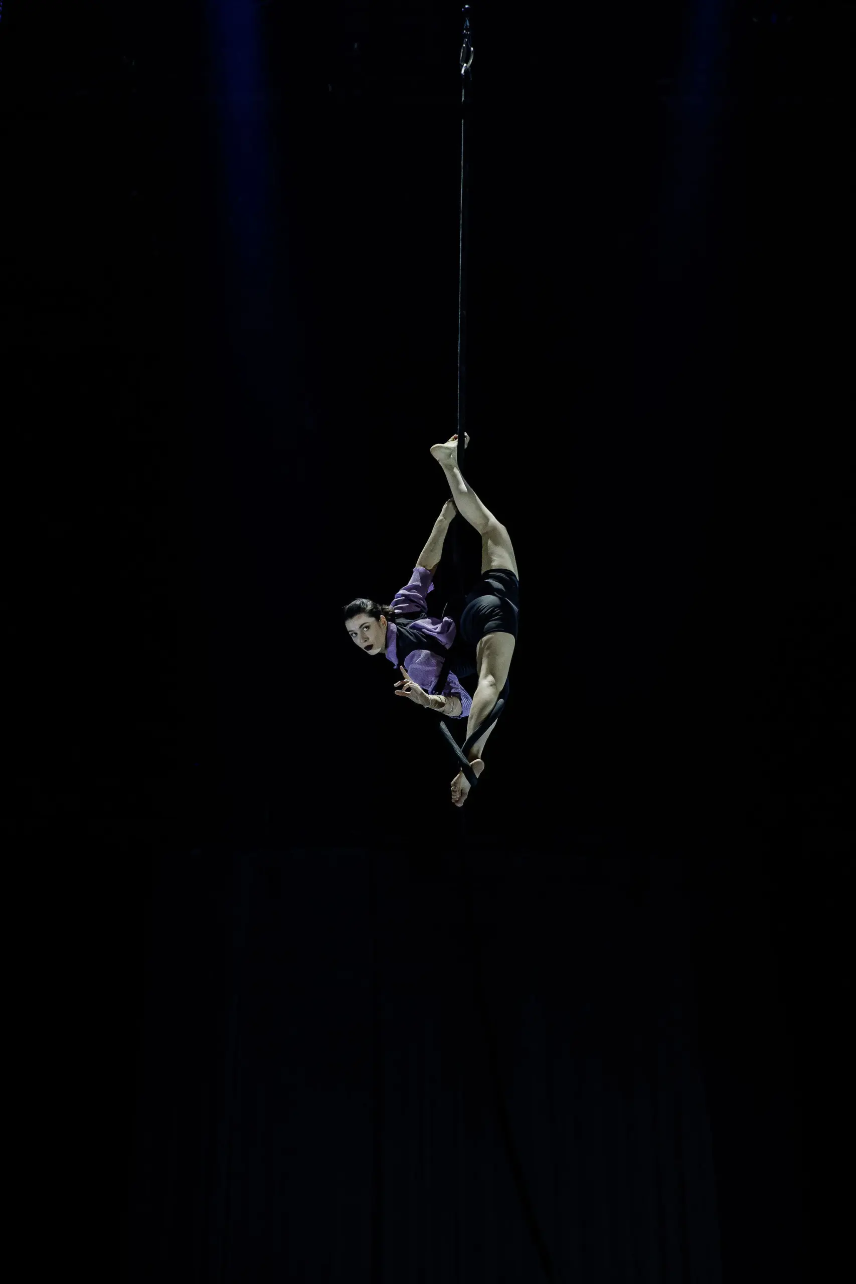 L'artiste aérienne Katarina Sobinkovicova se produit en hauteur, suspendue par une jambe à une corde, vêtue d'un haut violet et d'un short noir, sur un fond sombre.
