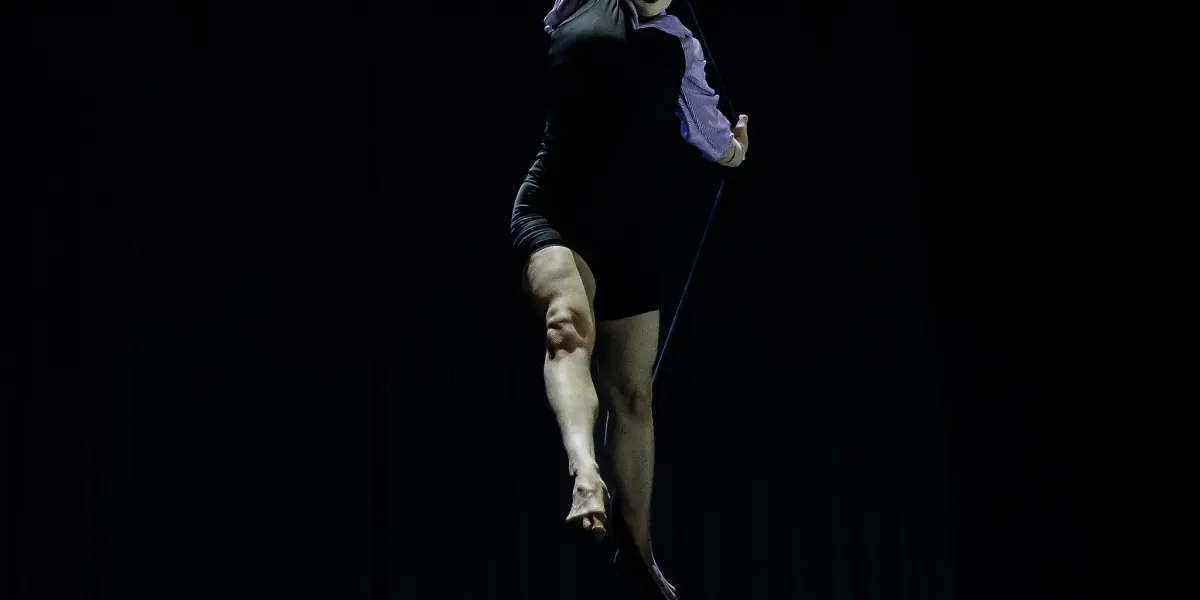 Katarina Sobinkovicova, vêtue de noir avec des manches violettes, exécute un numéro aérien, suspendue par un bras à une corde sur un fond sombre, ses jambes s'étendant gracieusement en dessous.