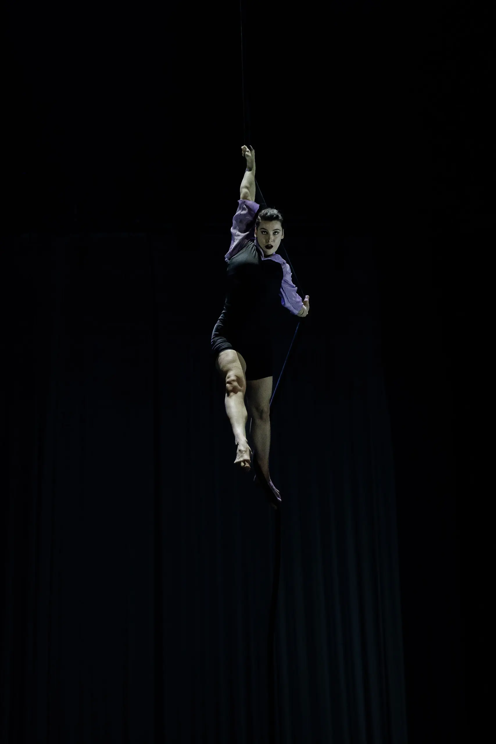 Katarina Sobinkovicova, vêtue de noir avec des manches violettes, exécute un numéro aérien, suspendue par un bras à une corde sur un fond sombre, ses jambes s'étendant gracieusement en dessous.
