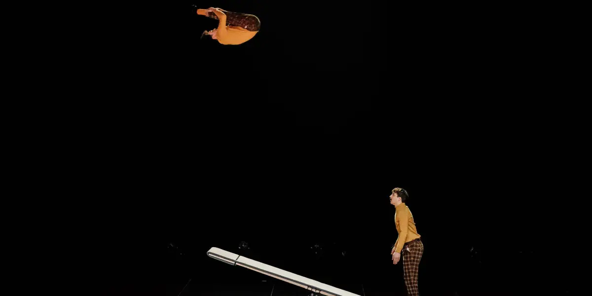 Léonard Meylan et Nathan Sterchi s'exécutent, l'un se lançant dans un saut périlleux au-dessus d'un appareil ressemblant à une balançoire, tandis que l'autre se tient sur le tapis en dessous, tous deux sur un fond sombre.