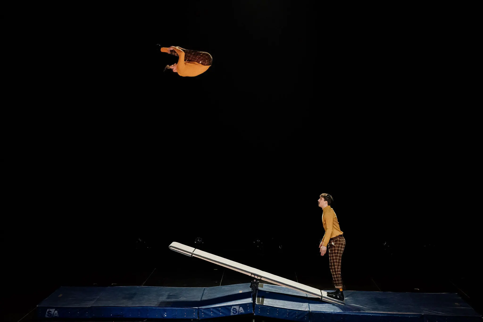 Léonard Meylan et Nathan Sterchi s'exécutent, l'un se lançant dans un saut périlleux au-dessus d'un appareil ressemblant à une balançoire, tandis que l'autre se tient sur le tapis en dessous, tous deux sur un fond sombre.