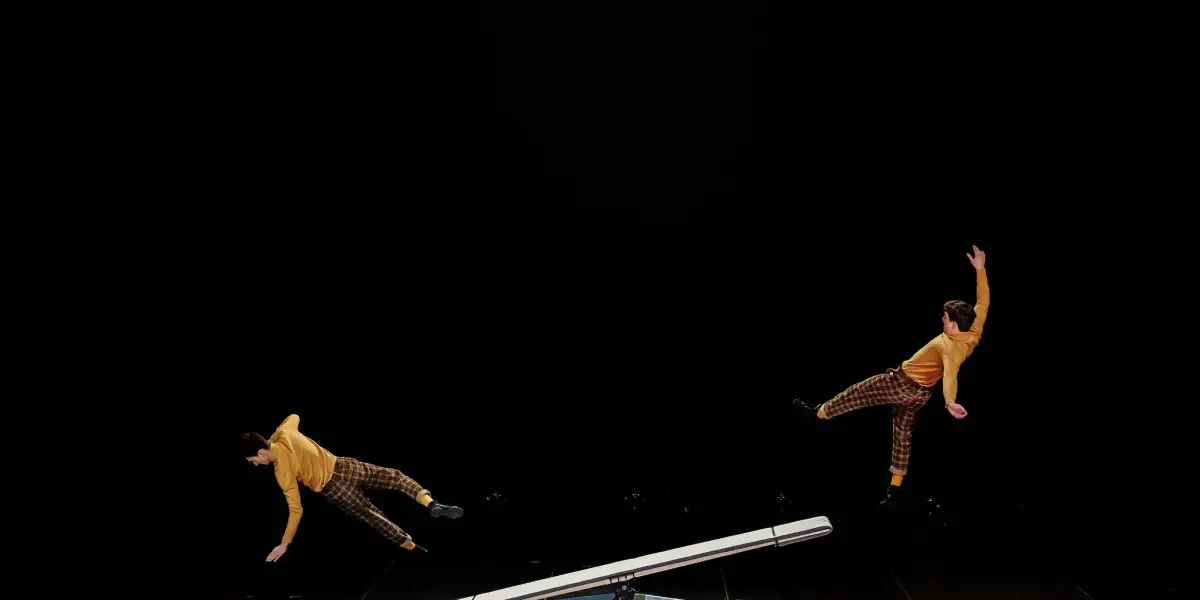 Léonard Meylan et Nathan Sterchi, deux acrobates en chemise jaune et pantalon à carreaux, sont en plein vol au-dessus d'une bascule sur des tapis bleus, réalisant une acrobatie sur fond noir.