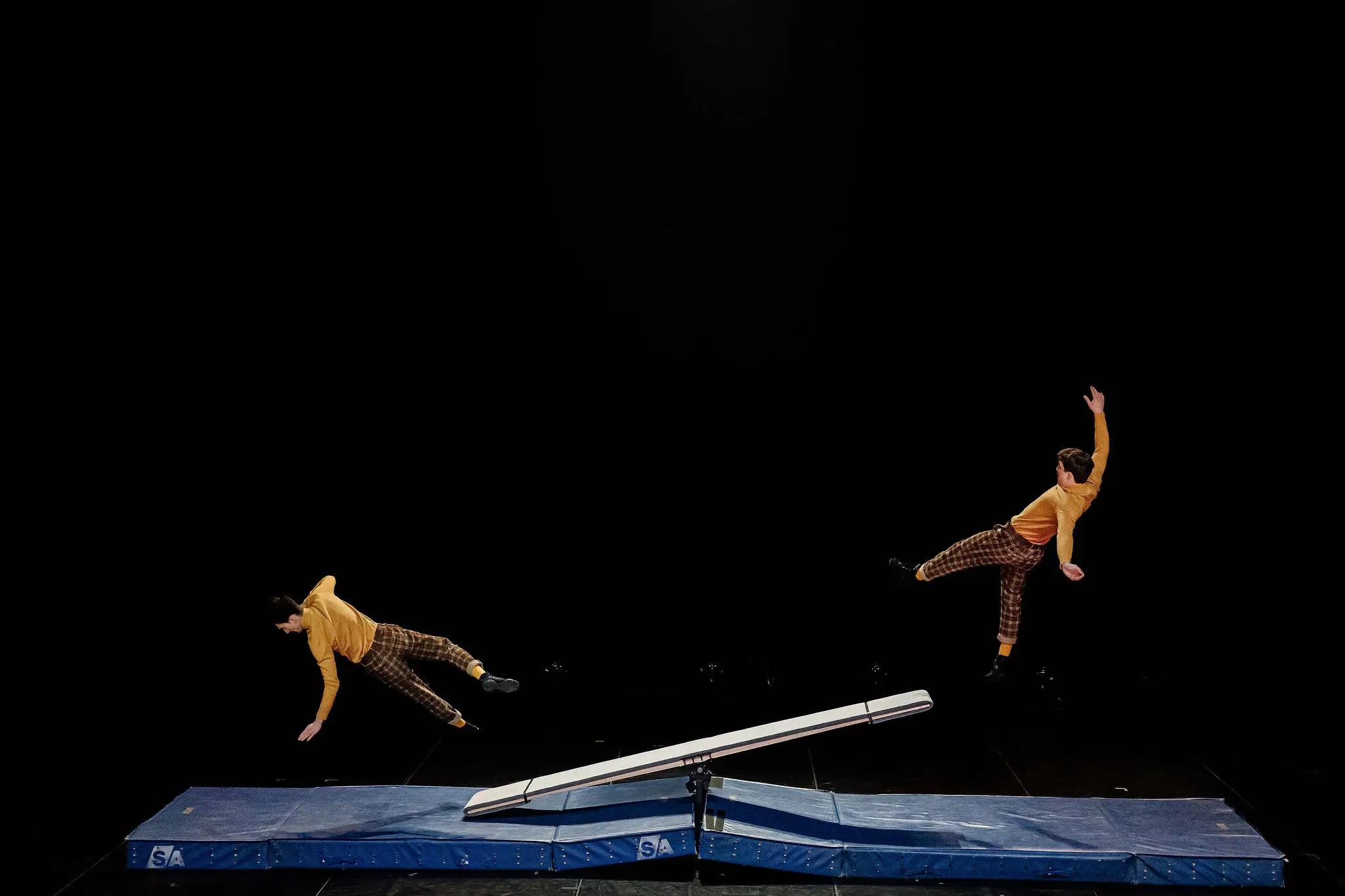 Léonard Meylan et Nathan Sterchi, deux acrobates en chemise jaune et pantalon à carreaux, sont en plein vol au-dessus d'une bascule sur des tapis bleus, réalisant une acrobatie sur fond noir.