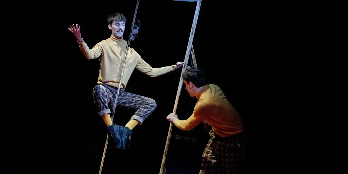 Deux performeurs, Léonard Meylan et Nathan Sterchi, vêtus de pulls jaunes et de pantalons écossais, interagissent avec un grand cadre rectangulaire vide. L'un est assis à l'intérieur du cadre, faisant des gestes, tandis que l'autre le tient et l'incline sur une scène sombre.