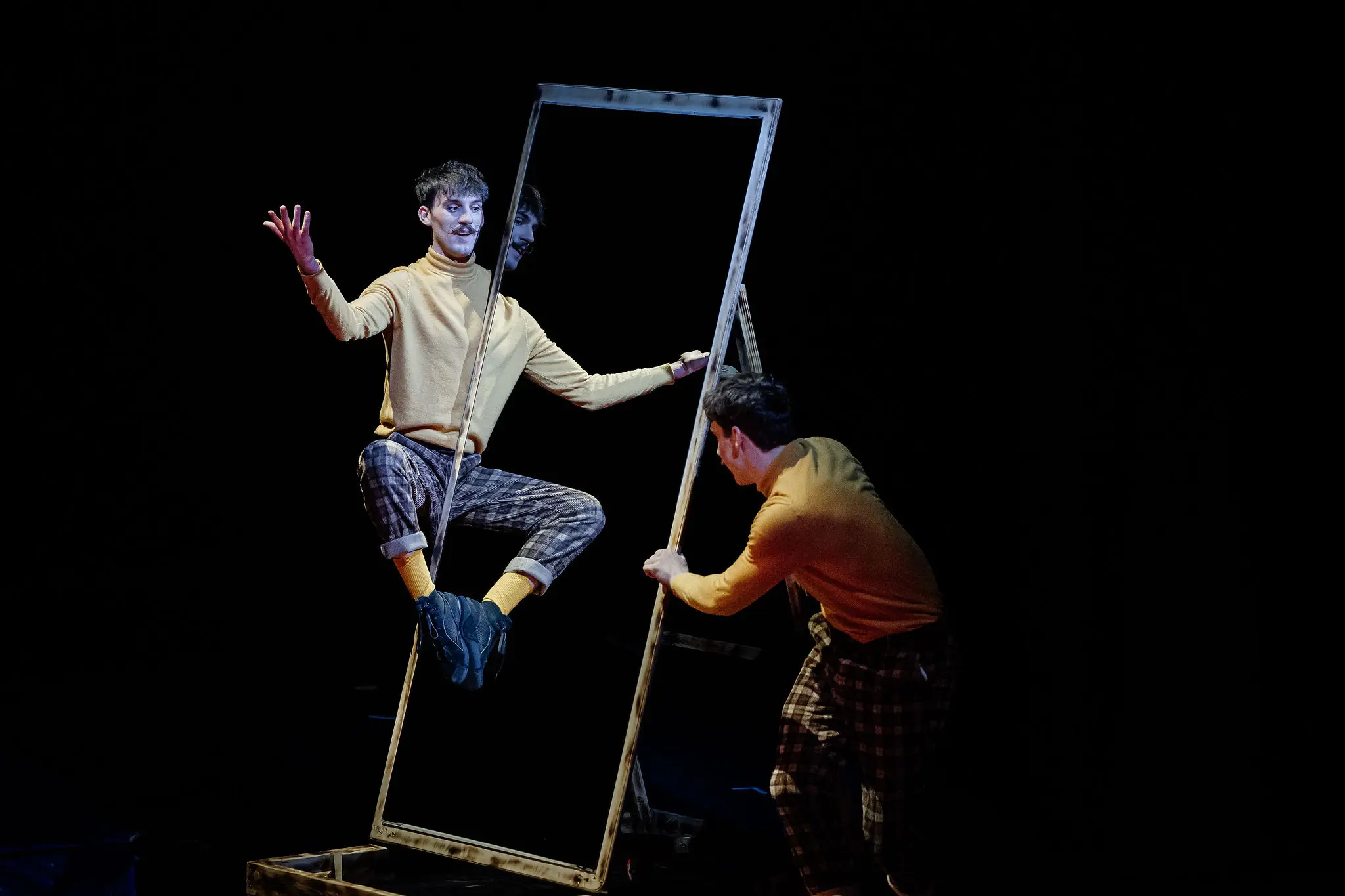 Deux performeurs, Léonard Meylan et Nathan Sterchi, vêtus de pulls jaunes et de pantalons écossais, interagissent avec un grand cadre rectangulaire vide. L'un est assis à l'intérieur du cadre, faisant des gestes, tandis que l'autre le tient et l'incline sur une scène sombre.