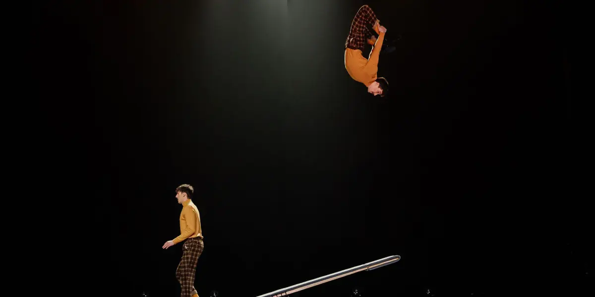 Deux acrobates, Léonard Meylan et Nathan Sterchi, vêtus de chemises jaunes et de pantalons à carreaux assortis, évoluent sur une balançoire à bascule ; l'un se tient au sol tandis que l'autre est en l'air, la tête en bas, dans un flip sur fond sombre.