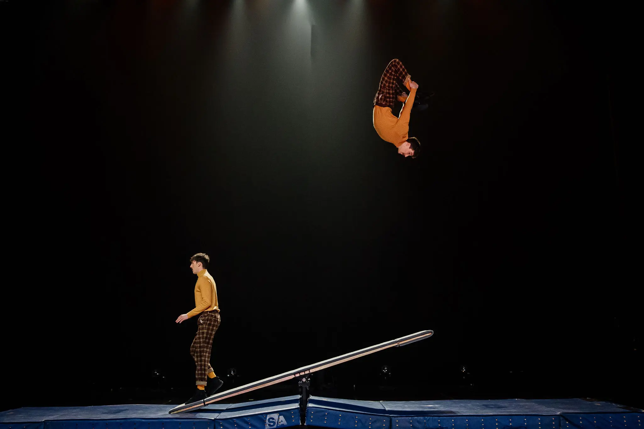 Deux acrobates, Léonard Meylan et Nathan Sterchi, vêtus de chemises jaunes et de pantalons à carreaux assortis, évoluent sur une balançoire à bascule ; l'un se tient au sol tandis que l'autre est en l'air, la tête en bas, dans un flip sur fond sombre.