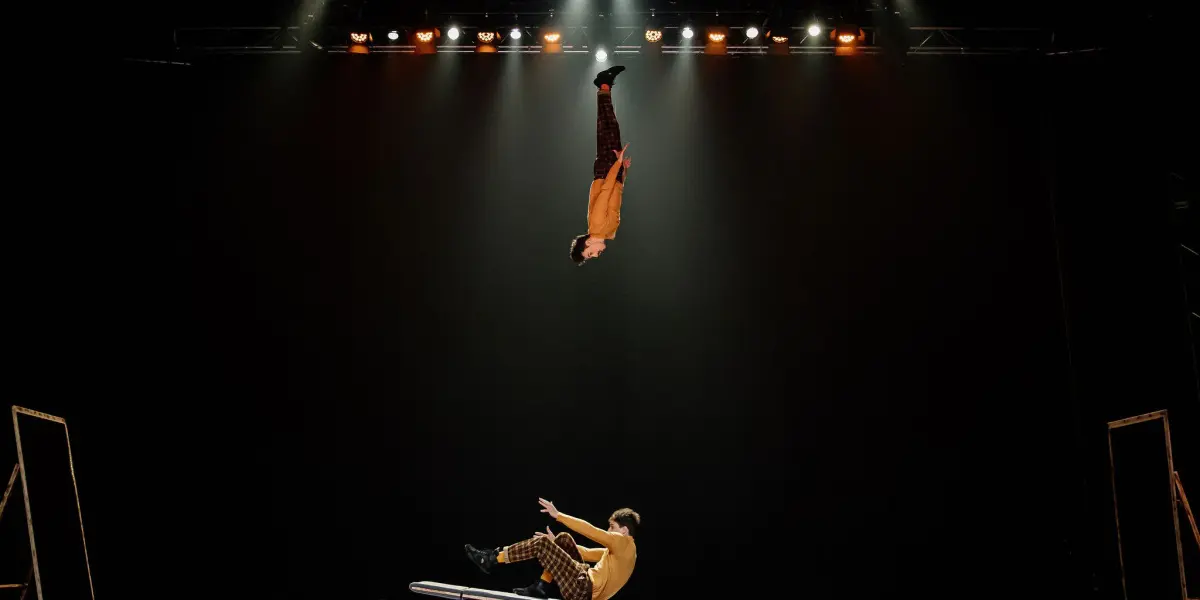 Deux acrobates, Léonard Meylan et Nathan Sterchi, se produisent sur scène ; l'un s'élève au-dessus d'une bascule tandis que l'autre tombe, tous deux vêtus de pantalons à motifs, au milieu de lumières vives sur un fond sombre.