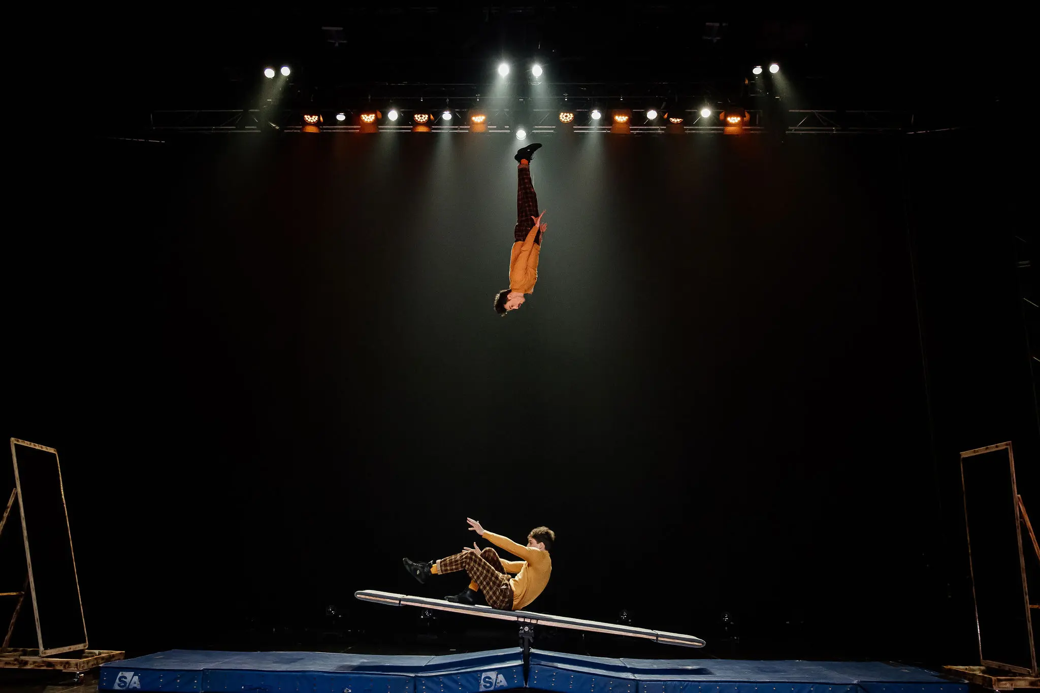 Deux acrobates, Léonard Meylan et Nathan Sterchi, se produisent sur scène ; l'un s'élève au-dessus d'une bascule tandis que l'autre tombe, tous deux vêtus de pantalons à motifs, au milieu de lumières vives sur un fond sombre.